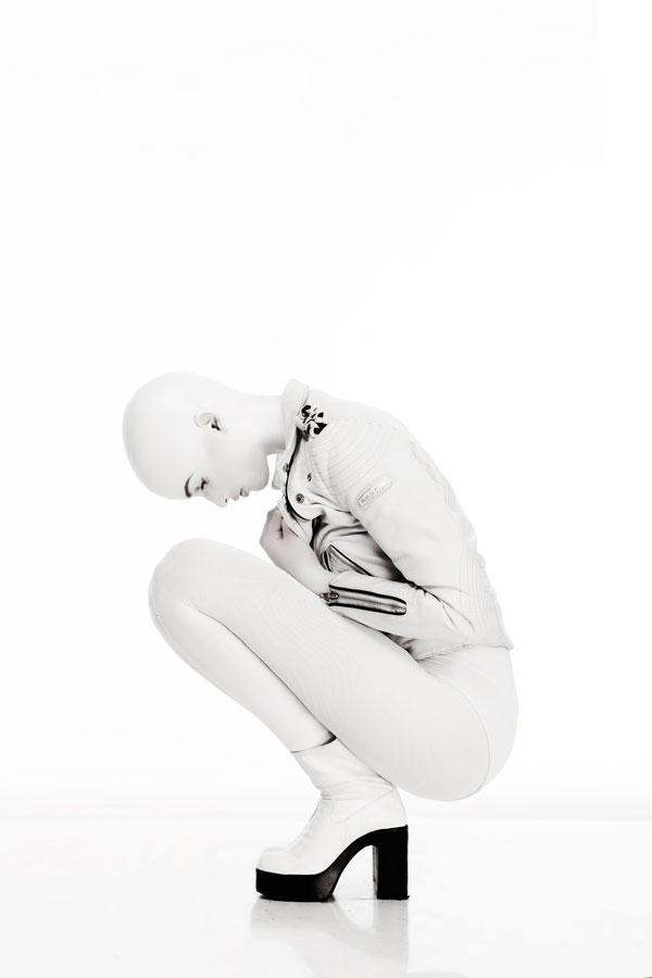Editorialfoto af model med skaldet hoved i helhvidt outfit med læderjakke og platformstøvler, sammenkrøben i hug mod ren hvid baggrund i minimalistisk futuristisk stil.