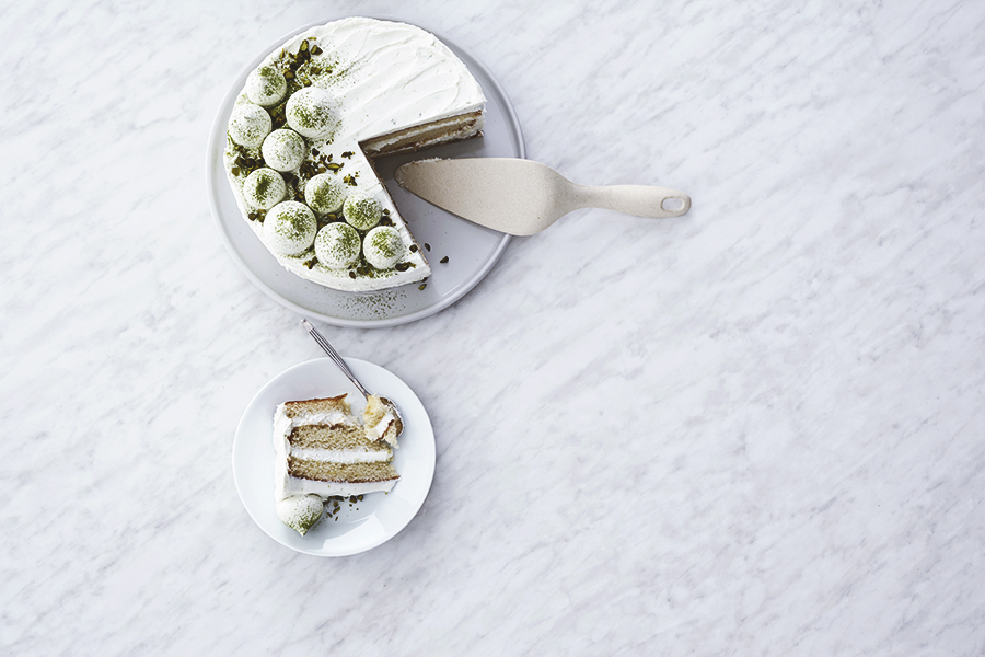 Lagkage med hvid creme og limepyntede kugler drysset med pistacie, fotograferet ovenfra på hvid marmor med et udskåret stykke på sidetallerken.