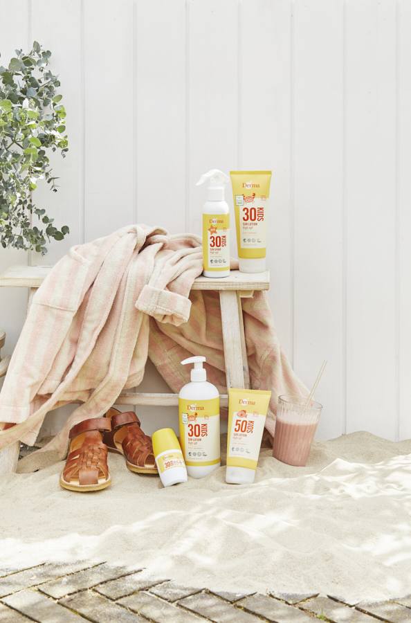 Still life af Derma Kids solcremeprodukter stylet på strand med sand, flettet lædersandal, lyserødt ternet håndklæde og smoothie foran hvid bræddevæg i sommerligt dagslys.